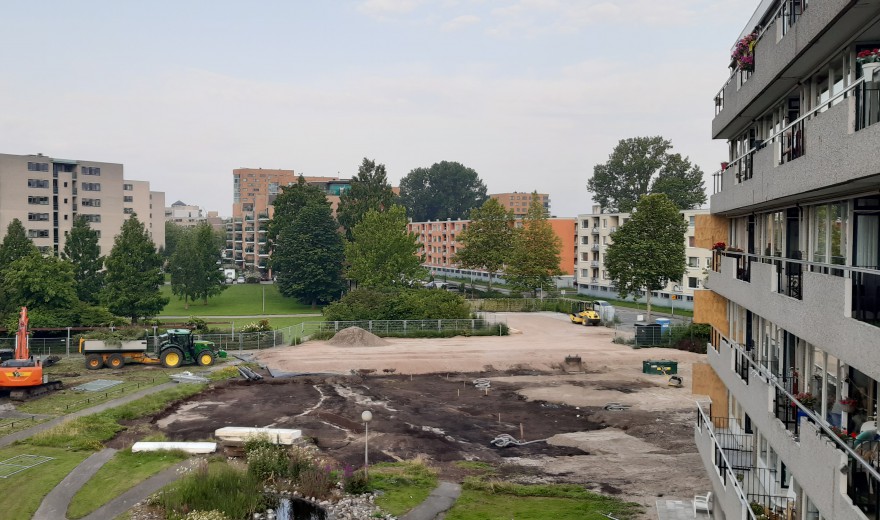Bouwplaats met graafmachines en zand, omringd door groene bomen en appartementencomplexen onder een bewolkte hemel.