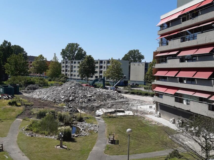 Afbeelding van een bouwplaats met puin naast een flatgebouw met rode zonneschermen, omgeven door groen en bomen.