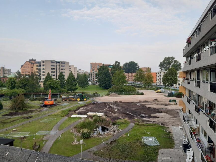 Luchtfoto van een stedelijk gebied met appartementen, een bouwplaats en graafmachines, omringd door groen en bomen.