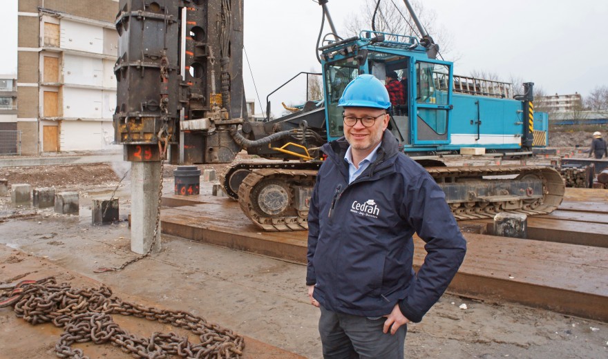 Man met blauwe helm staat voor een bouwplaats met zware machines en kettingen, klaar voor funderingswerkzaamheden.