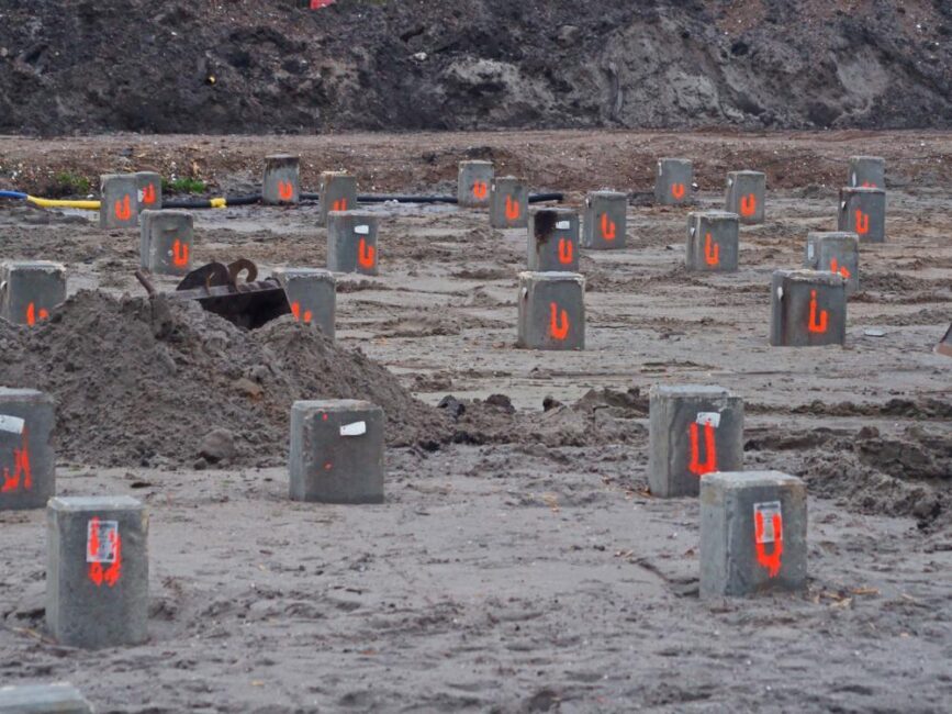 Bouwterrein met meerdere betonnen funderingspalen in de grond, genummerd met rode verf, omgeven door zand en aarde.