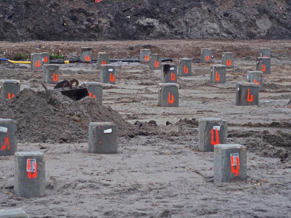 Bouwterrein met meerdere betonnen funderingspalen in de grond, genummerd met rode verf, omgeven door zand en aarde.