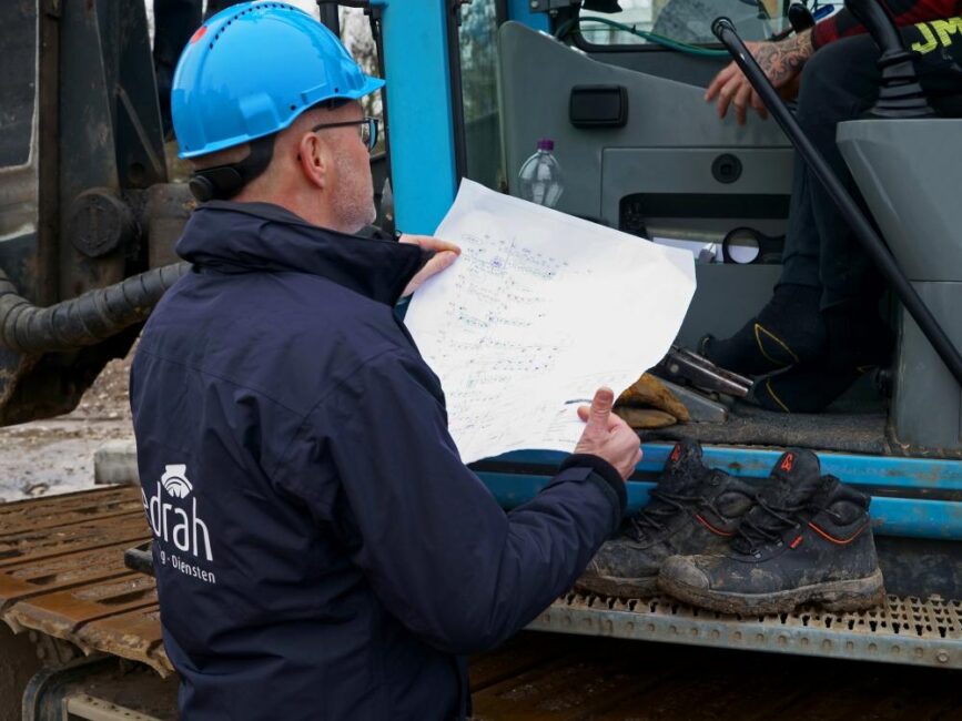 Man met blauwe helm en Cedrah-jas bekijkt bouwtekening naast graafmachine. Werkschoenen liggen op de machine.