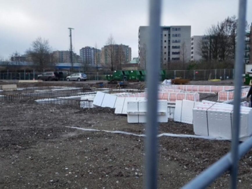 Bouwplaats met stapels witte isolatiematerialen, omheind met hekwerk. Op de achtergrond zijn hoge gebouwen zichtbaar.