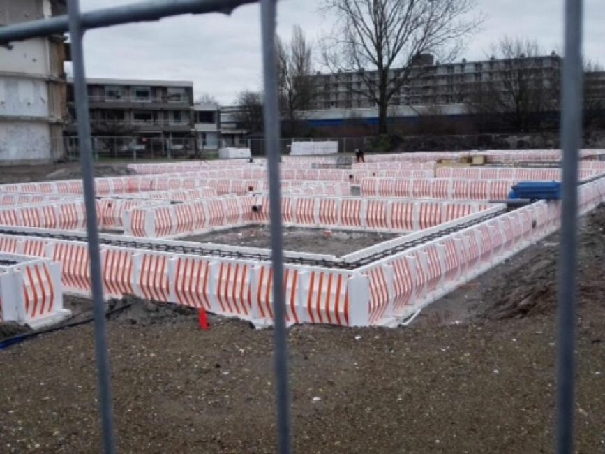Bouwplaats met fundering in aanbouw, omgeven door rood-witte veiligheidshekken. Op de achtergrond zijn gebouwen zichtbaar.