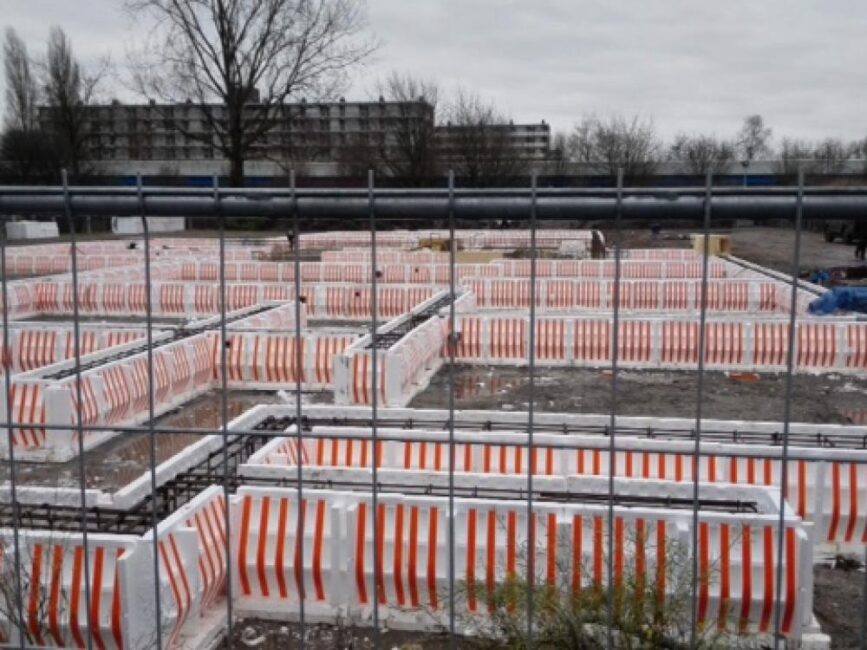 Bouwplaats met funderingen omgeven door oranje-witte afzetlinten, omheind door een hek. Op de achtergrond staan bomen.