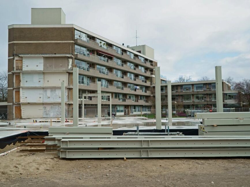 Bouwplaats met een gedeeltelijk gesloopt gebouw op de achtergrond en stapels bouwmaterialen op de voorgrond onder een bewolkte hemel.