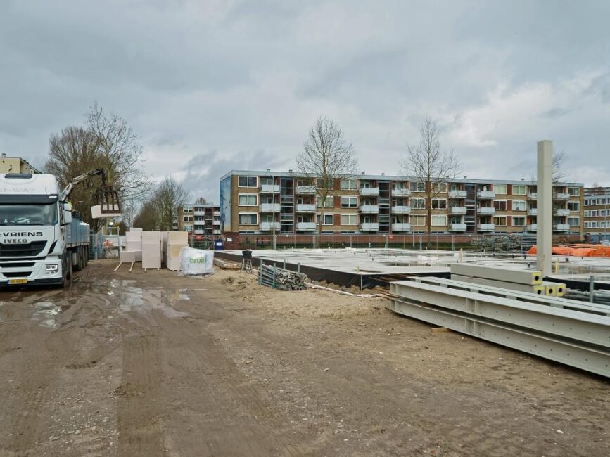 Bouwplaats met vrachtwagen en bouwmaterialen op de voorgrond, en een rij appartementen op de achtergrond onder een bewolkte hemel.