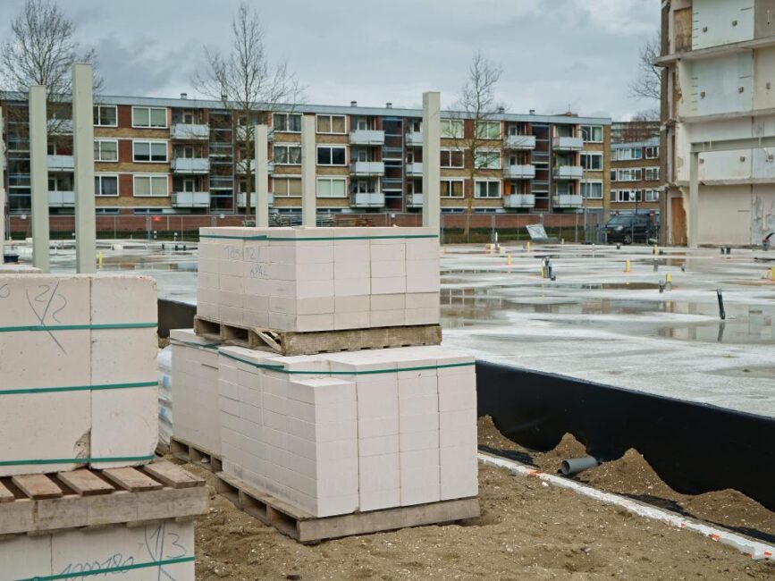 Bouwplaats met gestapelde witte bouwblokken op pallets. Op de achtergrond zijn appartementen en kale bomen zichtbaar.