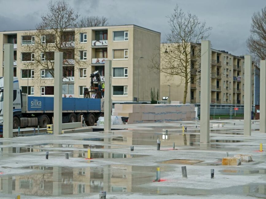 Bouwplaats met betonnen fundering en pilaren in opbouw. Op de achtergrond zijn woongebouwen en een vrachtwagen te zien.