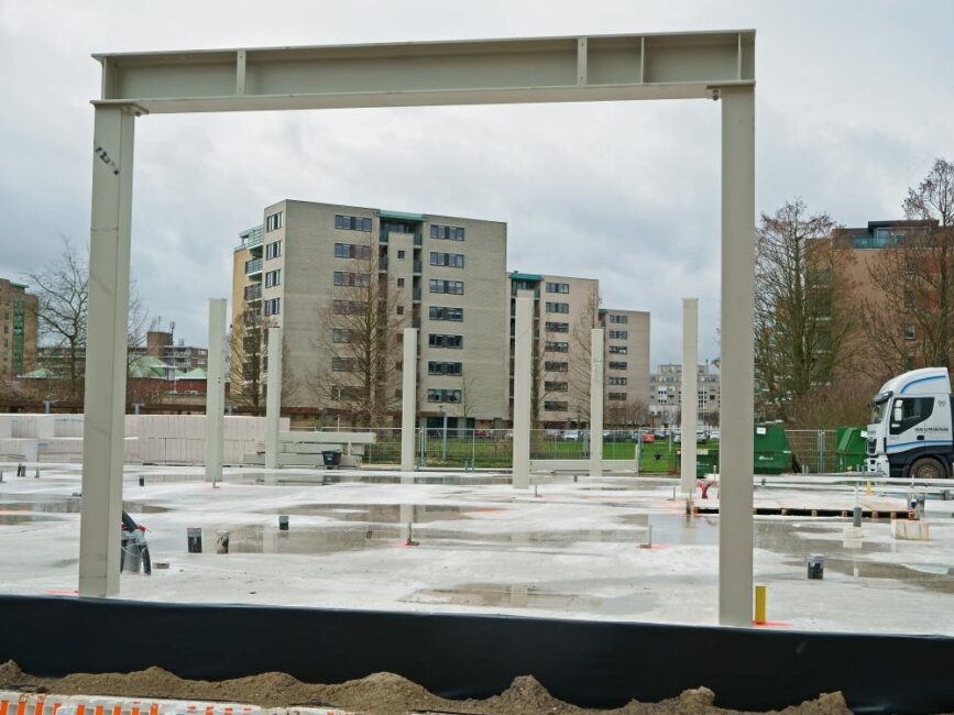 Bouwplaats met stalen frame in de voorgrond en hoge woongebouwen op de achtergrond onder een bewolkte hemel.