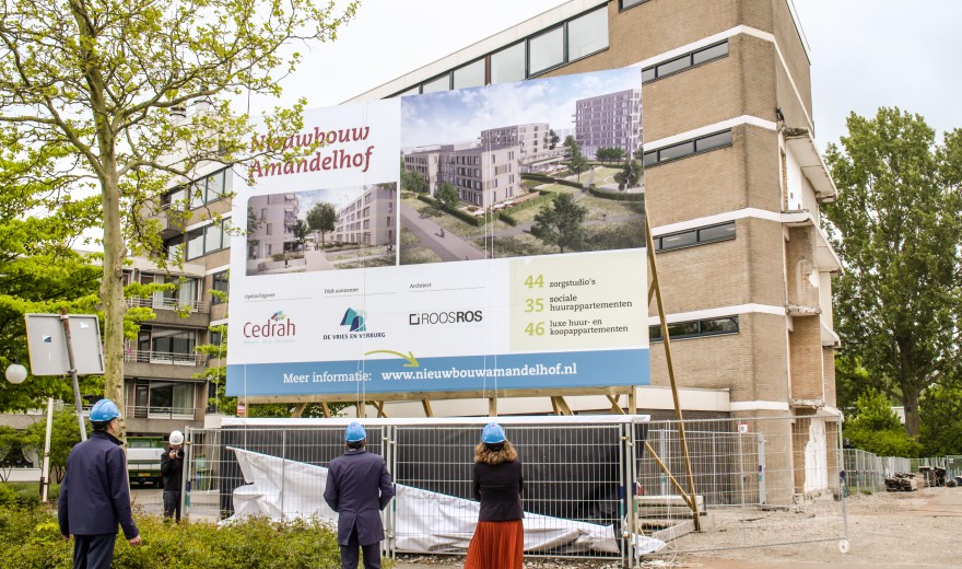 Drie mensen met bouwhelmen bekijken een bouwbord voor een nieuwbouwproject bij een bestaand gebouw in een groene omgeving.