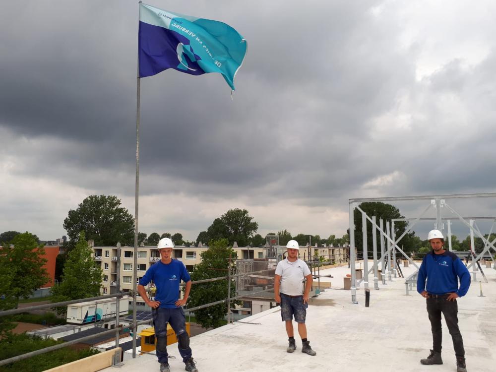 Drie bouwvakkers met helmen staan op een dak naast een wapperende vlag. De lucht is bewolkt en er zijn bomen op de achtergrond.