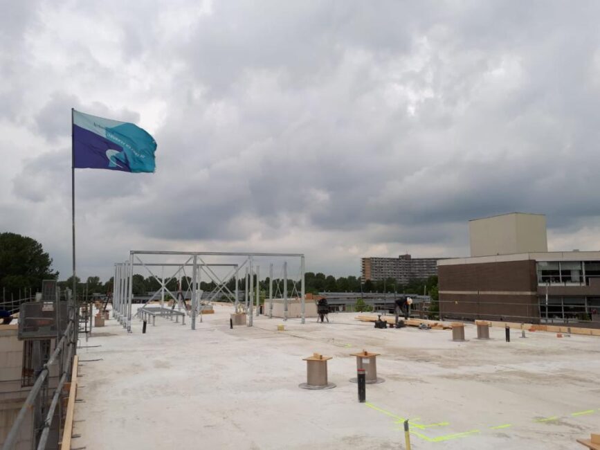 Vlag wappert op een dak met bewolkte lucht. Er zijn bouwmaterialen en een gebouw op de achtergrond zichtbaar.