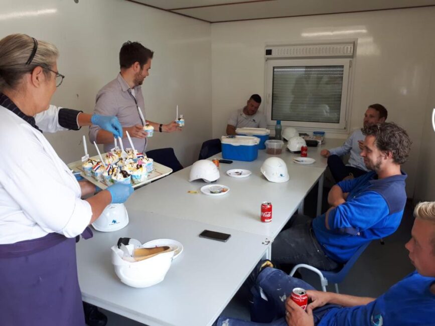 Een groep mensen zit aan een tafel in een kantine. Een vrouw serveert ijsjes op een dienblad. Veiligheidshelmen liggen op tafel.