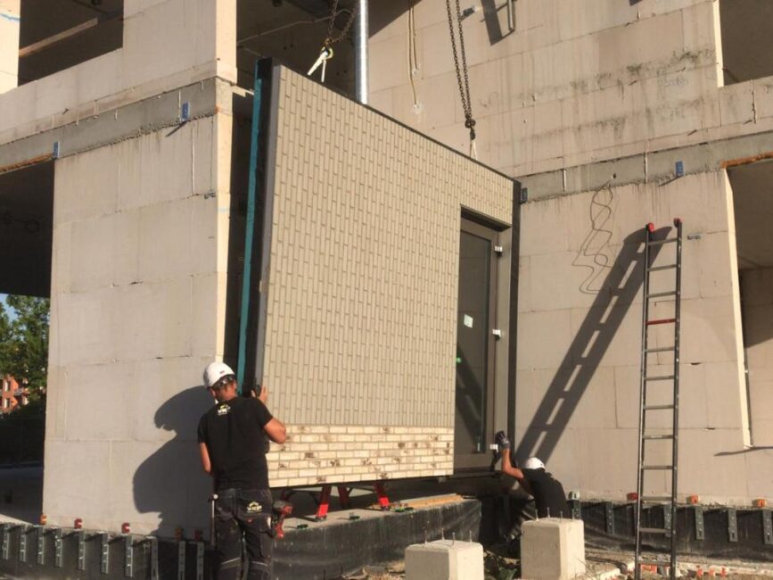 Twee bouwvakkers installeren een prefab gevelpaneel aan een gebouw. Een ladder staat tegen de muur. Het is zonnig weer.