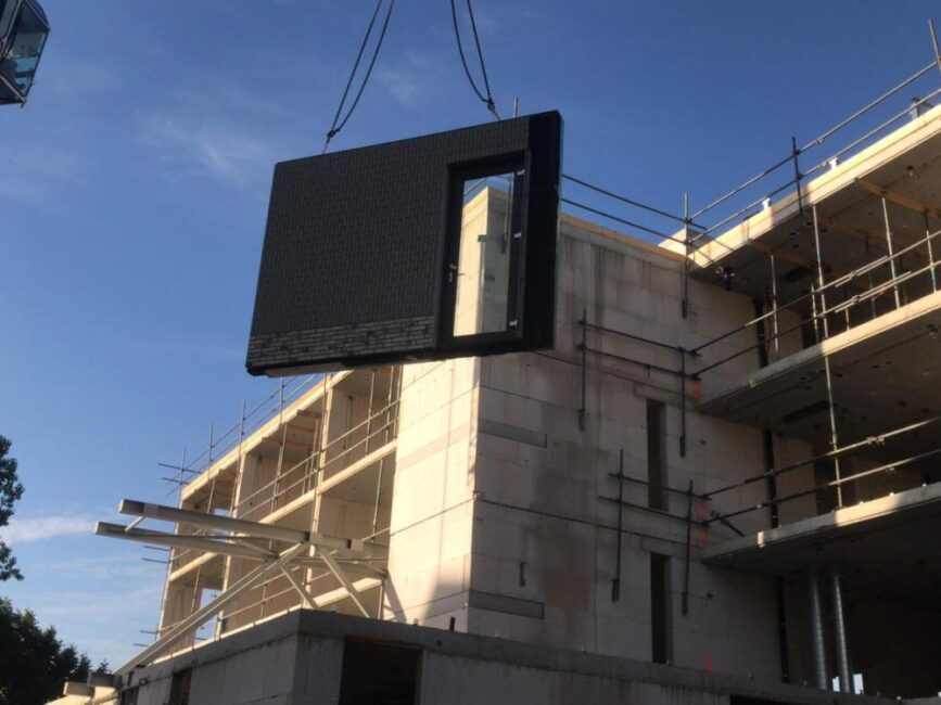 Een prefab betonnen muur met een raamopening wordt met een kraan opgetild bij een bouwplaats onder een blauwe hemel.