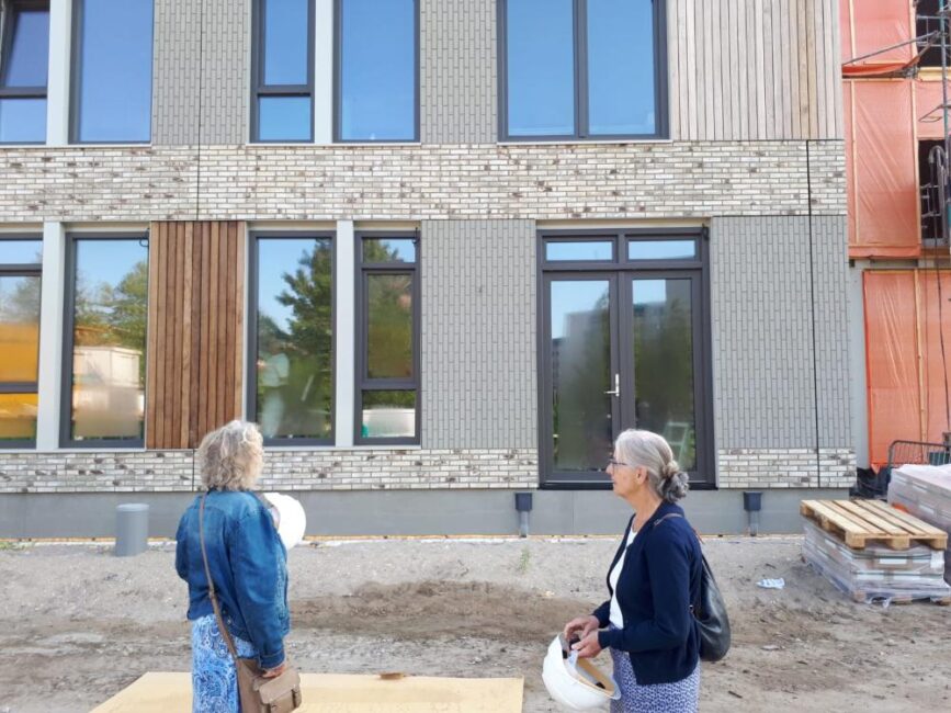 Twee mensen staan voor een modern gebouw in aanbouw, met bakstenen en houten panelen. Ze dragen bouwhelmen in hun handen.