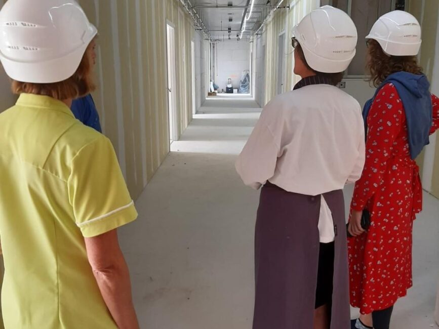 Drie mensen met helmen bekijken een gang in aanbouw. Ze dragen verschillende kledingstijlen en staan met hun rug naar de camera.