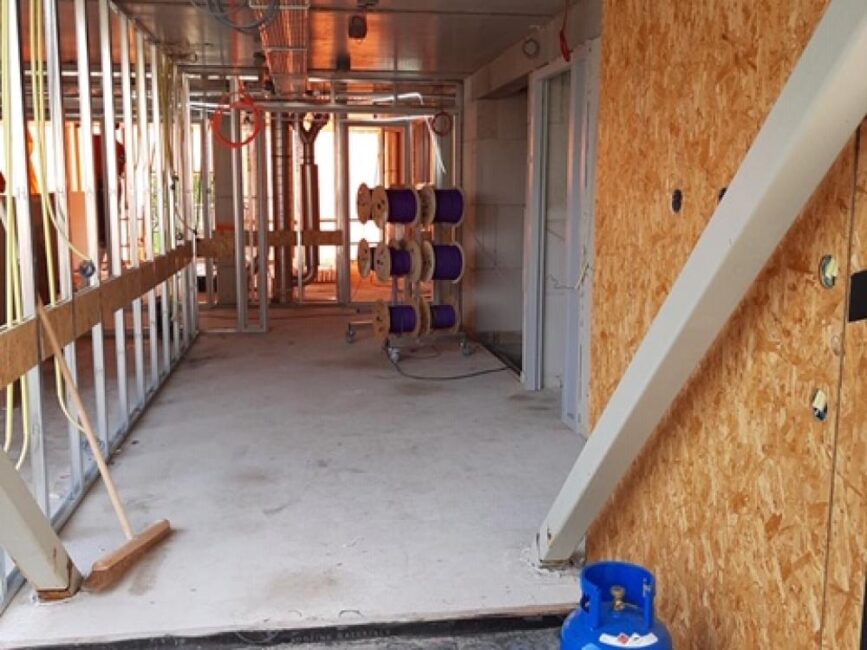 Bouwplaats met onafgewerkte muren, leidingen en een blauwe gasfles. Er zijn houten panelen en kabelrollen zichtbaar.