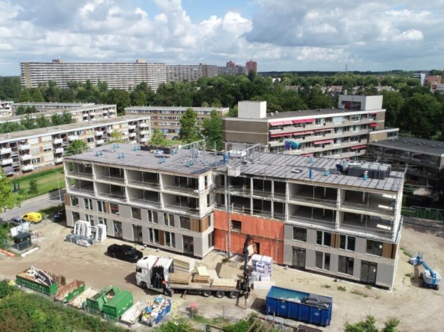 Luchtfoto van een bouwplaats met een bijna voltooid appartementencomplex, omringd door groen en andere gebouwen onder een blauwe hemel.