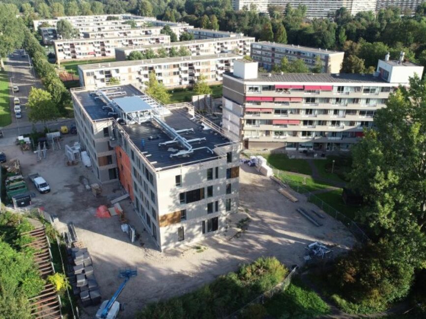 Luchtfoto van een bouwplaats met een bijna voltooid gebouw, omringd door groen en bestaande wooncomplexen in een stedelijk gebied.