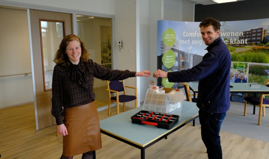 Twee mensen schudden handen in een kantoorruimte. Op de tafel staat een doos met vakjes en een banner op de achtergrond.