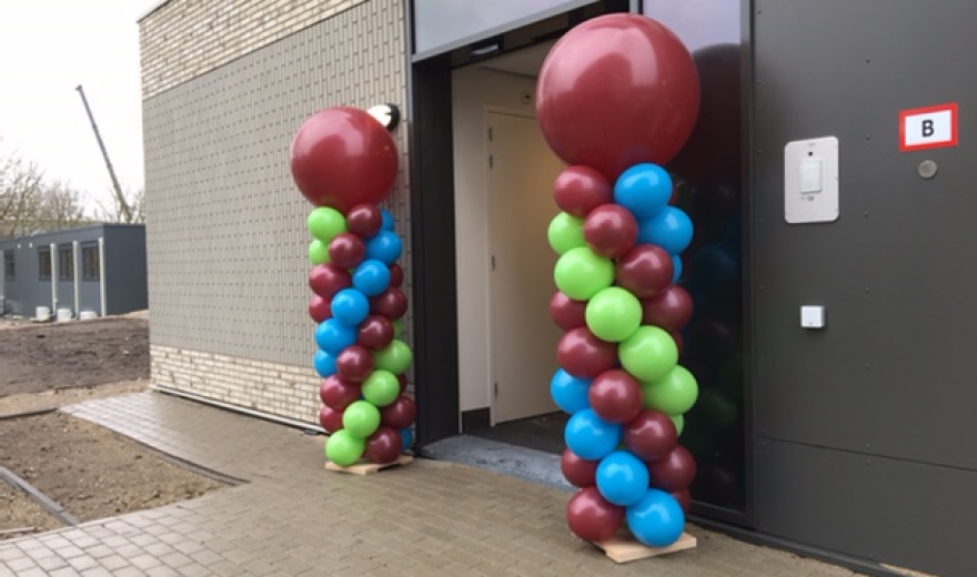 Twee kleurrijke ballonpilaren met rode, blauwe en groene ballonnen staan bij de ingang van een modern gebouw met bakstenen muur.