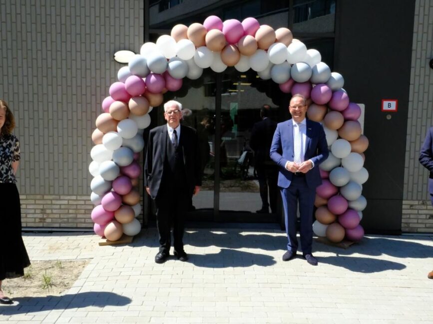 Twee mannen in pakken staan voor een boog van gekleurde ballonnen bij de ingang van een gebouw, met mensen op de achtergrond.