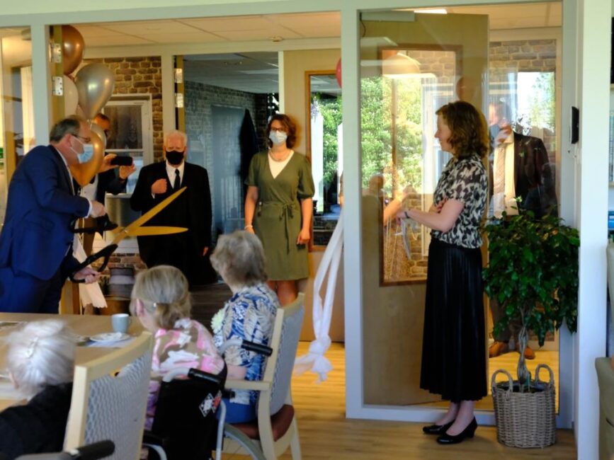 Een man knipt een lint door met een grote schaar tijdens een ceremonie. Omstanders dragen mondkapjes en kijken toe.