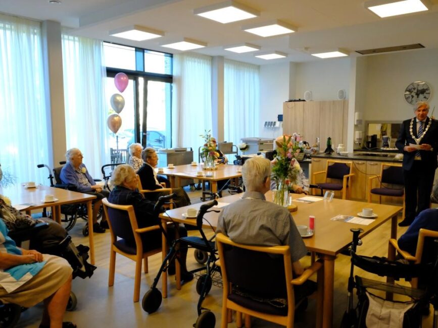 Een groep ouderen zit in een gemeenschappelijke ruimte met ballonnen en bloemen, terwijl een spreker voor hen staat.