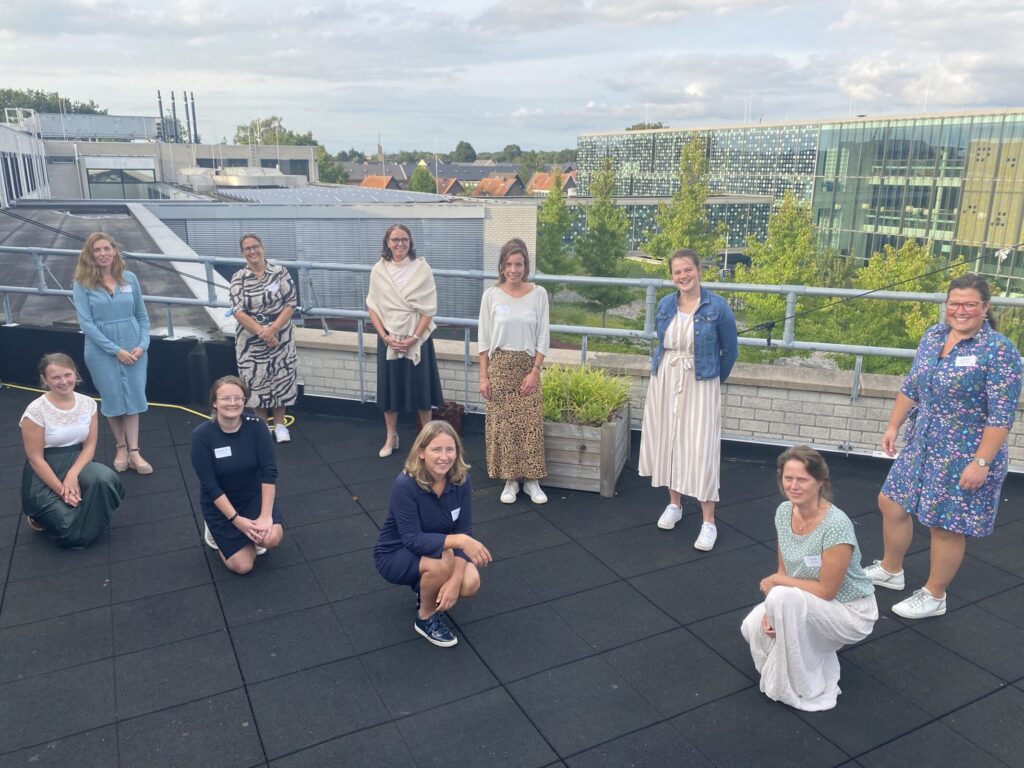 Groep mensen poseert op een dakterras met gebouwen en bomen op de achtergrond, onder een bewolkte hemel.