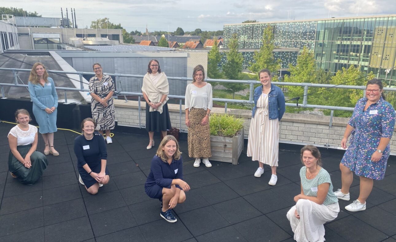 Groep mensen poseert op een dakterras met gebouwen en bomen op de achtergrond, onder een bewolkte hemel.