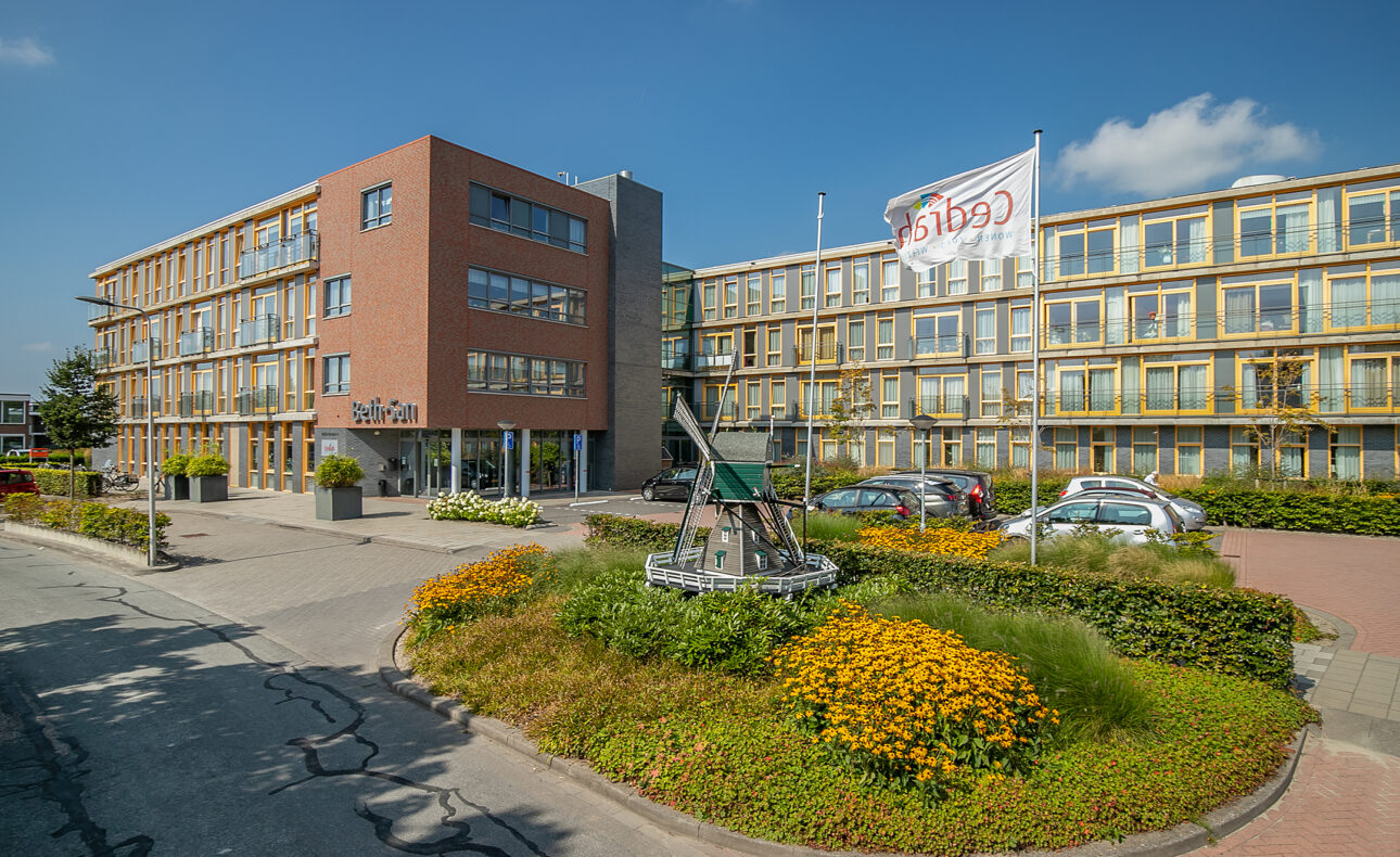 Vooraanzicht van een modern gebouw met meerdere verdiepingen, omgeven door een tuin met bloemen en een vlag met het logo van Cedrah.