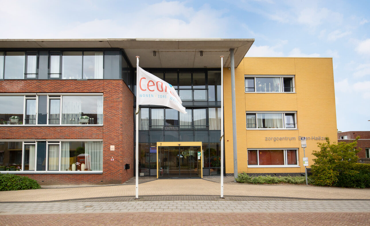 Vooraanzicht van een modern gebouw met een rode en gele gevel. Twee vlaggen wapperen voor de ingang onder een blauwe lucht.