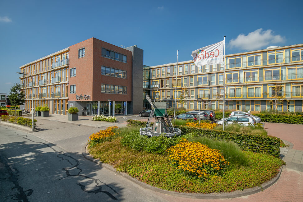 Vooraanzicht van een modern gebouw met meerdere verdiepingen, omgeven door een tuin met bloemen en een vlag met het logo van Cedrah.