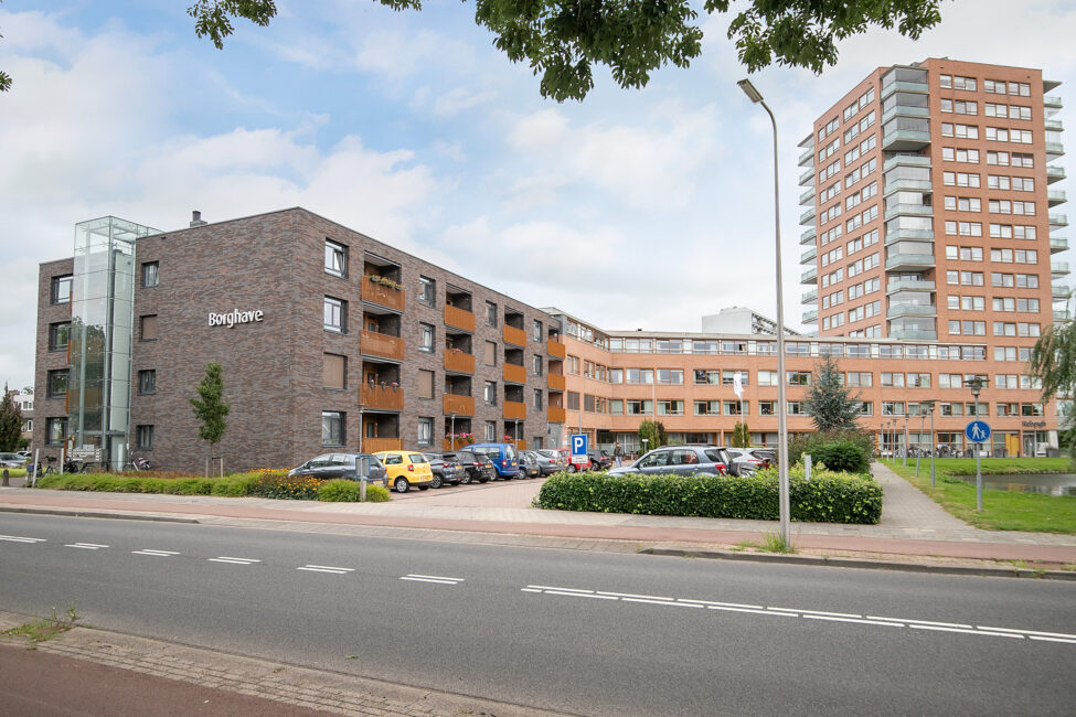 Appartementencomplex met meerdere verdiepingen, omgeven door geparkeerde auto's en groen, onder een bewolkte hemel.
