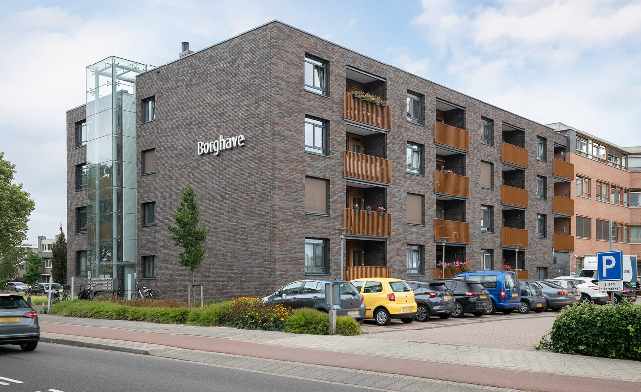 Afbeelding van een modern, vier verdiepingen tellend appartementencomplex met bakstenen gevel en geparkeerde auto's ervoor.