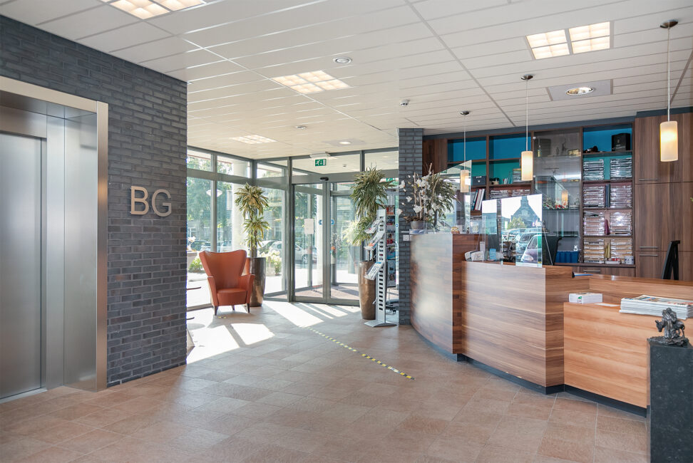 Ingang van een modern gebouw met een houten receptiebalie, glazen deuren, een lift en een oranje fauteuil bij het raam.