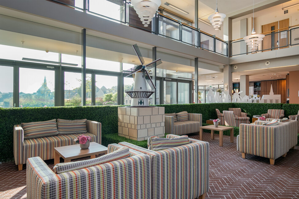 Zonnige lounge met gestreepte banken, houten tafels en een decoratieve molen. Grote ramen bieden uitzicht op groene omgeving.