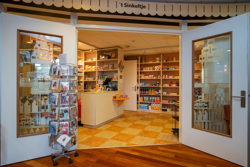 Open deuren naar een winkel met houten vloeren, een rek met ansichtkaarten links en planken vol producten aan de achterkant.
