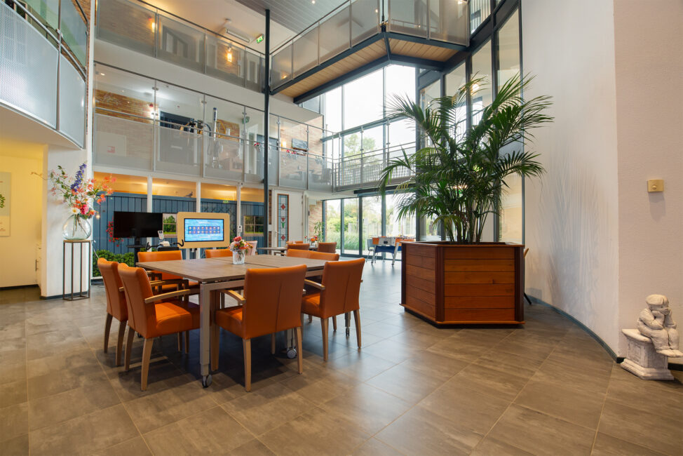 Interieur van een moderne ruimte met een grote tafel en oranje stoelen, omringd door glazen wanden en groene planten.