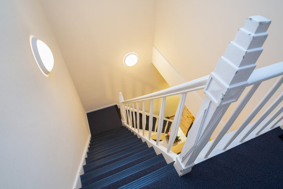 Trap met blauwe treden en witte leuningen, gezien van bovenaf. Twee ronde lampen verlichten de witte muren van het trappenhuis.