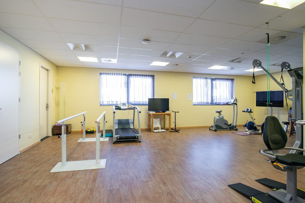 Een lichte fitnessruimte met loopband, hometrainer en andere fitnessapparatuur, houten vloer en ramen met jaloezieën.
