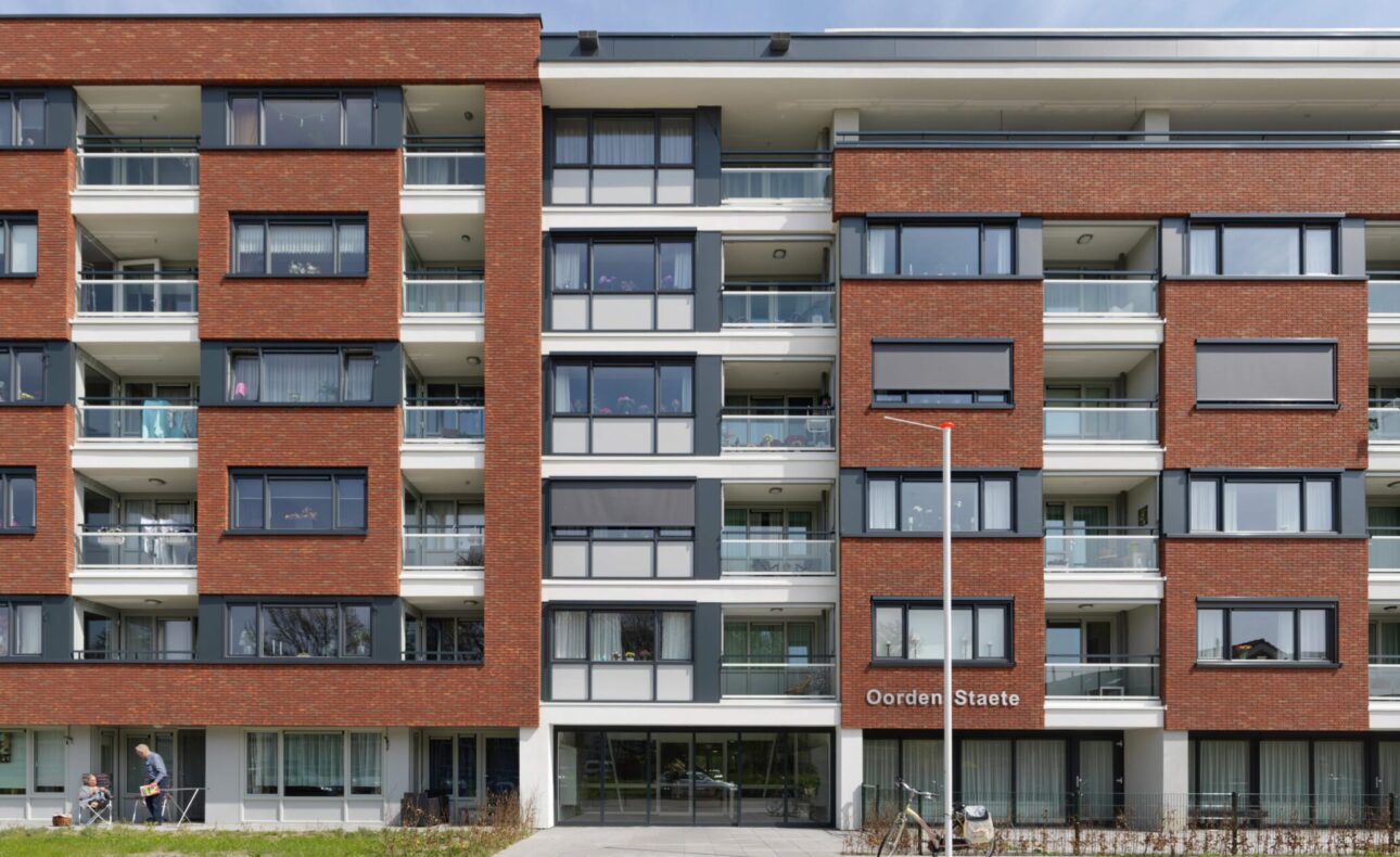 Afbeelding van een modern appartementencomplex met rode bakstenen en witte balkons, onder een heldere blauwe lucht.