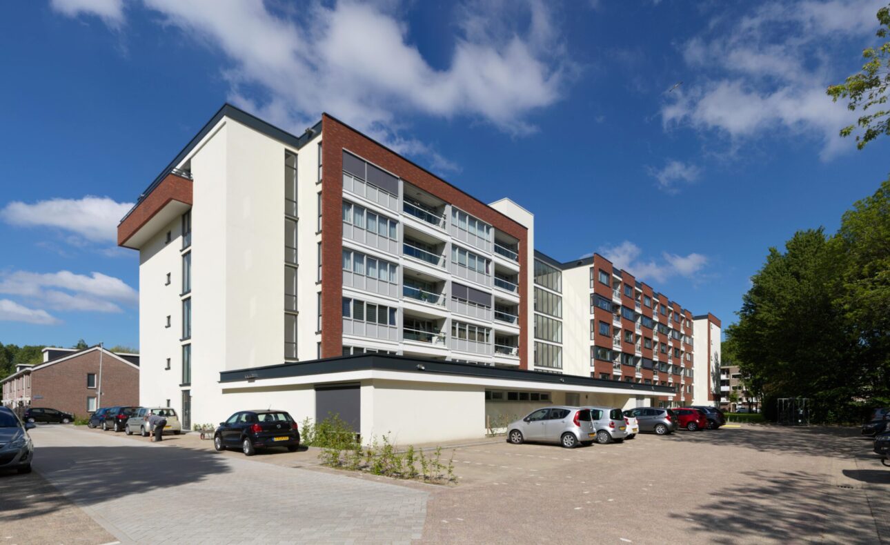 Afbeelding van een modern, meerlagig appartementencomplex met balkons, geparkeerde auto's en een blauwe lucht met enkele wolken.