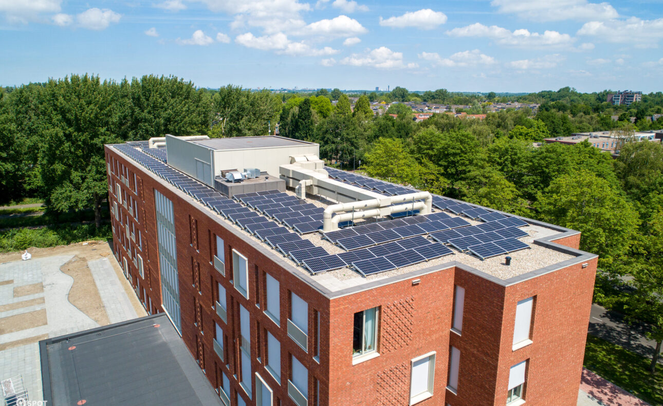 Luchtfoto van een modern rood bakstenen gebouw met zonnepanelen op het dak, omgeven door groene bomen en een blauwe lucht.