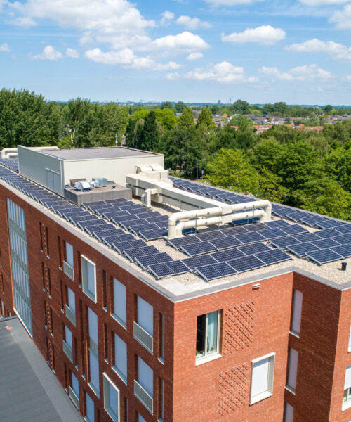 Luchtfoto van een modern rood bakstenen gebouw met zonnepanelen op het dak, omgeven door groene bomen en een blauwe lucht.