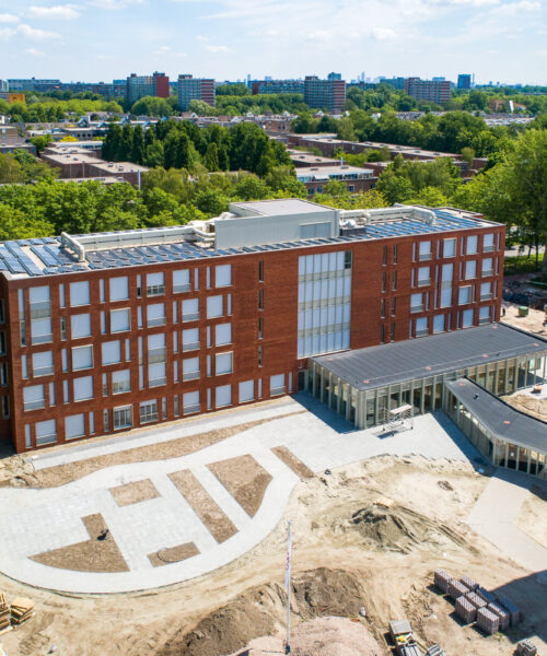 Luchtfoto van een modern, rood bakstenen gebouw omgeven door groen en stedelijke omgeving op een zonnige dag in Vlaardingen.