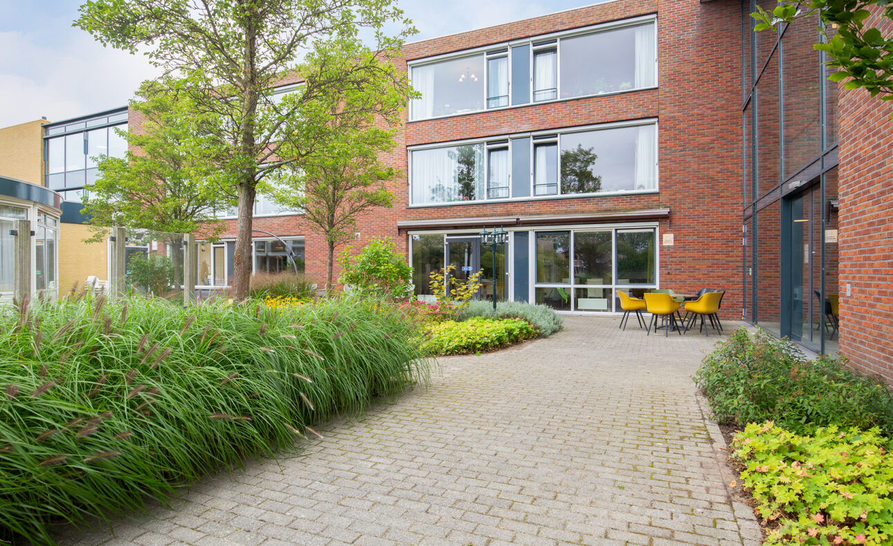 Buitenaanzicht van een modern gebouw met rode bakstenen, grote ramen en een tuin met groene planten en een terras met stoelen.